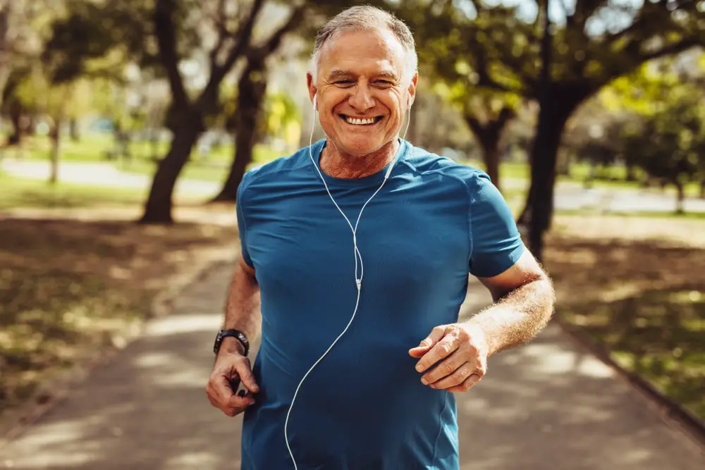 Senior-man-smiling-while-jogging-through-park-wearing-headphones