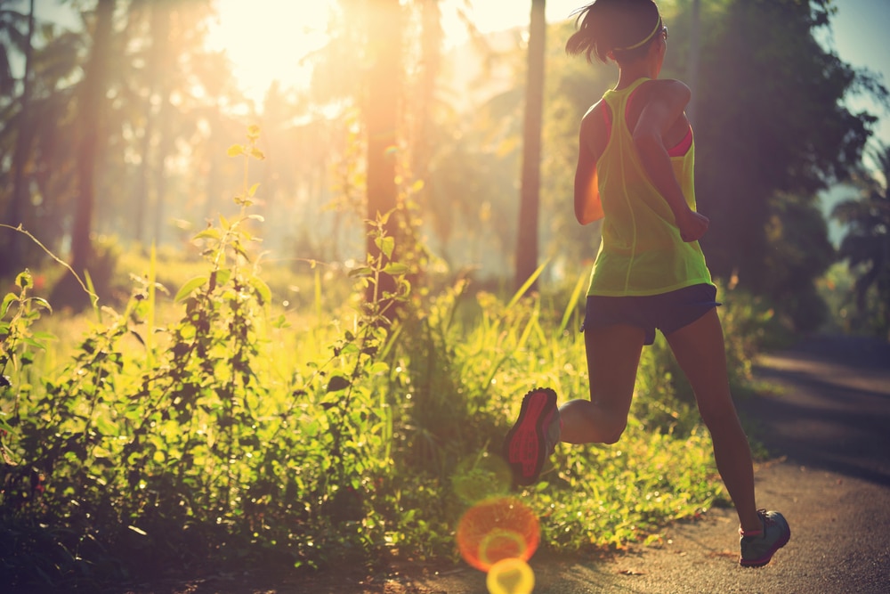 Young-fit-woman-running-along-path-in-forested-area-with-sun-shining-through-trees.